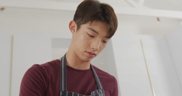 Young man concentrating while cooking in a modern kitchen. He is wearing a striped apron and casually dressed. Useful for illustrating cooking classes, home cooking concepts, culinary arts training, or modern lifestyle scenes.