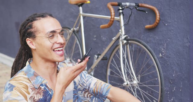 Young man with dreadlocks sitting outdoors using smartphone voice command, smiling cheerfully. Behind him is a modern bicycle with a purple background suggesting urban environment. Ideal for themes of technology in everyday life, relaxation, millennials, summer, transportation, urban living.