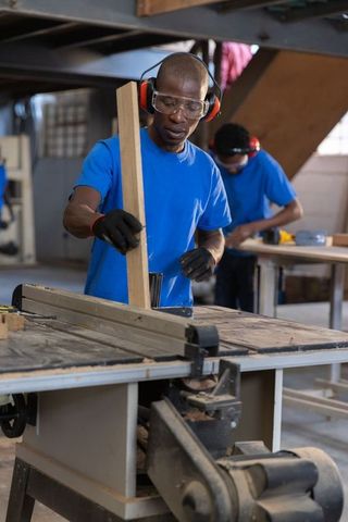 African American Carpenter Positioning Wood on Table Saw Wearing Safety Gear in Workshop