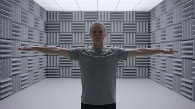 Woman standing with arms outstretched inside anechoic test chamber wearing gray tee
