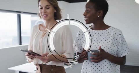 Two women holding tablet and mug discussing data with hud overlay in modern office