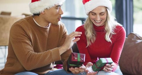 Couple exchanging christmas gifts on sofa wearing santa hats, smiling warmly