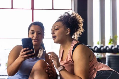 Diverse women sharing smartphone and reviewing workout progress in sunlit gym