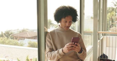 Mid-adult man leaning on glass door checking smartphone on sunlit balcony with greenery