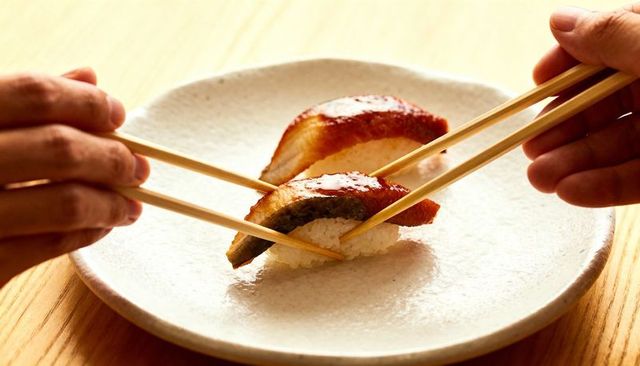 Grasping hands picking up glazed unagi nigiri sushi with chopsticks intimate closeup