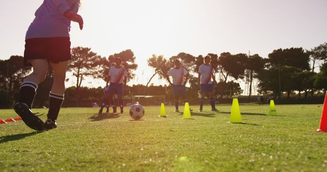 Group of young soccer players running drills and training on a grass field with cones. The scene is set during sunset, highlighting the athletic energy and teamwork. Suitable for use in sports promotion, youth development programs, or physical education materials.