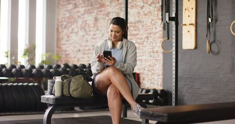 Asian woman sitting on gym bench checking smartphone wearing hoodie and headphones