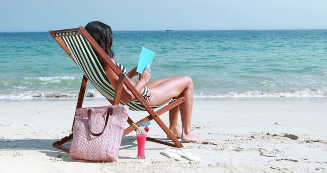 Woman sitting on a striped lounge chair on the sandy beach, enjoying a book with a turquoise cover. Beside her are a beach bag and a tropical drink. Perfect for travel blogs, vacation advertisements, relaxation promotion, or summer holiday ideas.