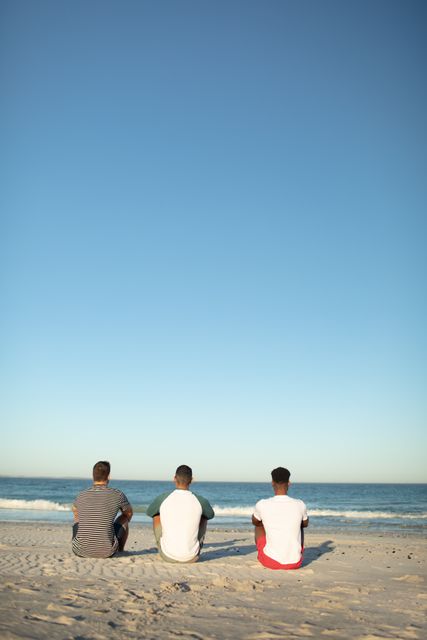 Three male friends are sitting on the sandy beach, facing the ocean under a clear blue sky. This image can be used for themes related to friendship, relaxation, summer vacations, and outdoor leisure activities. It is ideal for travel brochures, social media posts about beach outings, and advertisements promoting summer destinations.