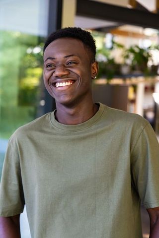 Smiling young black man wearing olive t-shirt by glass door with plant shelf, natural light