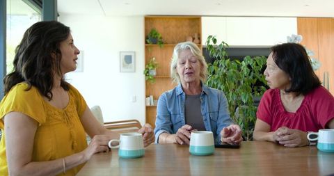 Diverse women sharing conversation and coffee around sunlit kitchen table with smartphone