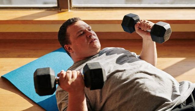 Overweight man lifting dumbbells on yoga mat during sunlit home workout