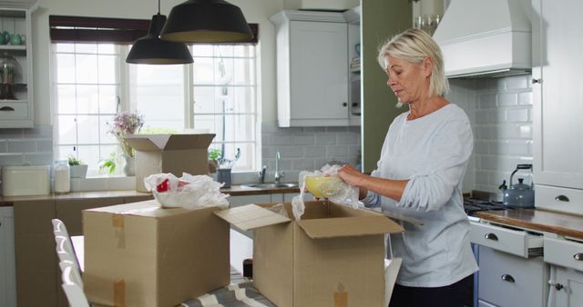 A senior woman is seen in a modern kitchen, surrounded by cardboard boxes, unpacking items. The image depicts the process of moving into a new home, suggesting themes of relocation, starting fresh, and domestic life. Ideal for use in articles/blogs about moving, retirement, lifestyle changes, and home organization and decoration.
