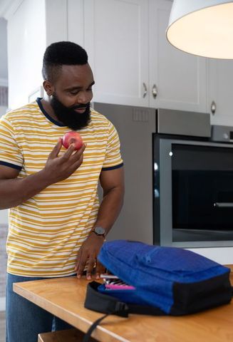 African american man packing blue backpack while holding apple at modern kitchen island