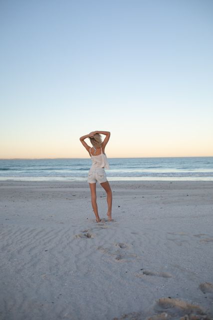 Woman standing with hands behind her head, facing the ocean, relaxing on a sandy beach during sunset. Perfect for use in travel advertisements, wellness campaigns, and lifestyle blogs promoting relaxation and tranquility.