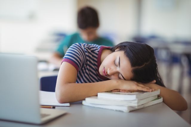 Young schoolgirl resting her head on books while sleeping in a classroom. Ideal for illustrating concepts of student fatigue, academic stress, need for rest, and challenges in education. Useful for educational articles, blogs about student life, and health-related content on sleep and productivity.