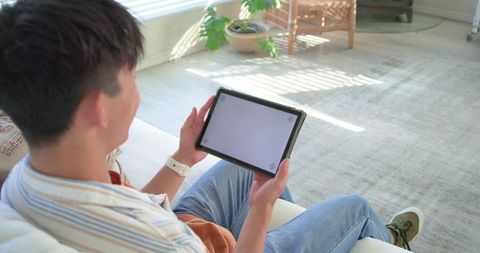 Asian man relaxing on sofa holding tablet with blank screen in sunlit modern living room