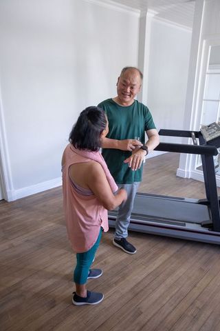 Chinese senior couple using smartwatch and treadmill for home cardio training, smiling