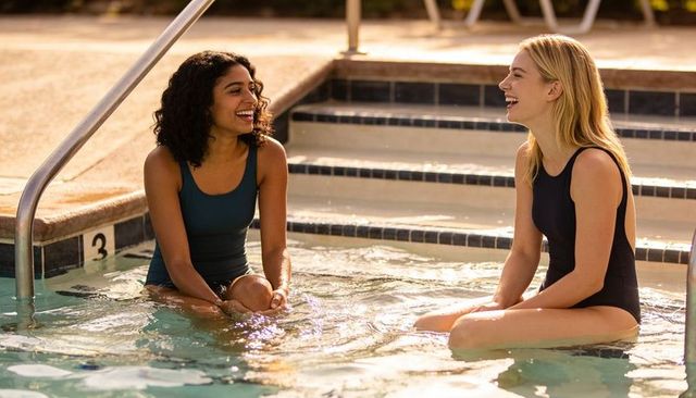 Laughing friends chatting on submerged pool steps in sunlit summer one-piece swimsuits