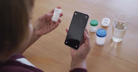 Mature woman checking daily medication with smartphone at home table with pill organizers
