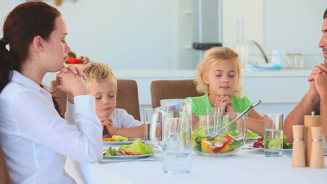 Family sitting together at dining table, saying prayers before eating. Mixed aged group with parents and children showing gratitude for the meal. Features a meal with fresh vegetables on table, revealing a harmonious family time. Useful for illustrating family bonding, cultural traditions, or lifestyle articles about the importance of mealtime conversations.