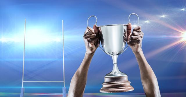 Hands of an athlete holding a trophy high in a stadium, symbolizing triumph and achievement. Rugby goalposts are visible in the background, adding context to the competitive nature of the event. Suitable for use in sports promotion, motivational content, advertisements, and articles highlighting success and victorious moments in athletics.