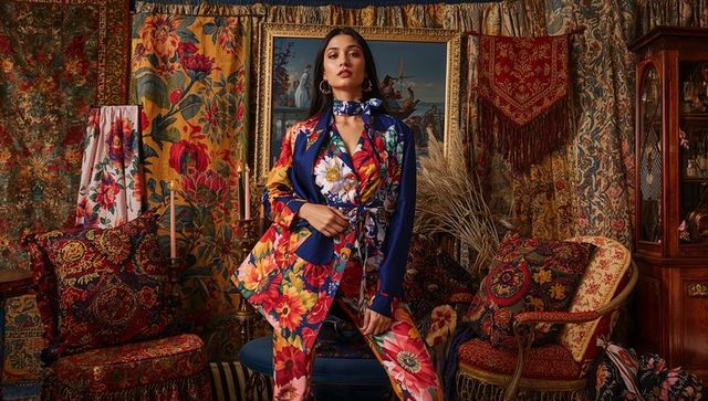 Model posing in vibrant floral suit and scarf among luxe maximalist vintage tapestries
