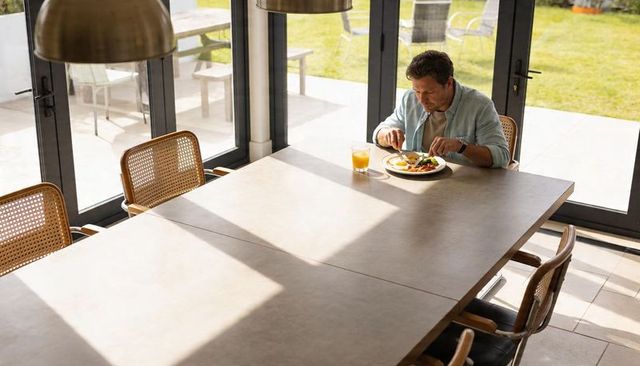 Solitary man eating breakfast at sunlit dining table by glass doors overlooking patio lawn