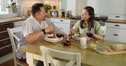 Asian couple enjoying cozy candlelit dinner at farmhouse kitchen table with wine