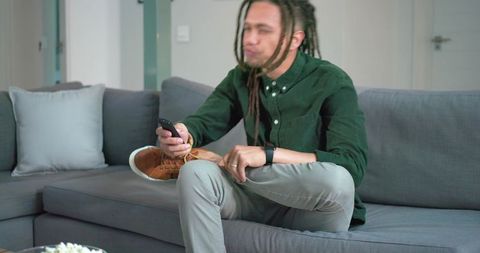 Man sitting on gray sofa adjusting shoe while holding remote in modern apartment living room