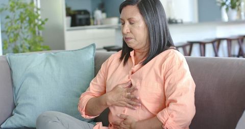 Mature woman sitting on couch experiencing chest and stomach discomfort in living room