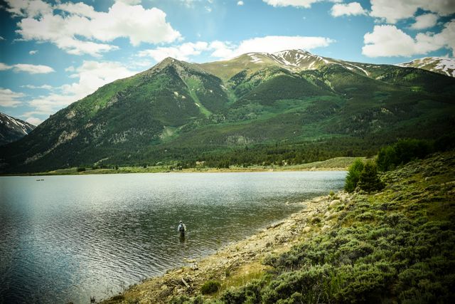 Beautiful scene depicting a lone fisherman standing in a tranquil alpine lake, surrounded by lush green mountains and under a sky dotted with clouds. Ideal for use in nature tourism promotions, outdoor activity advertisements, and travel blog visuals.