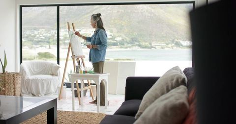 Senior woman painting on easel in sunlit waterfront living room wearing denim casual outfit