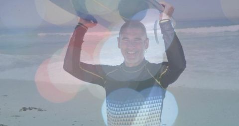 Smiling man wearing wetsuit holding flotation boards overhead on sandy beach with bokeh