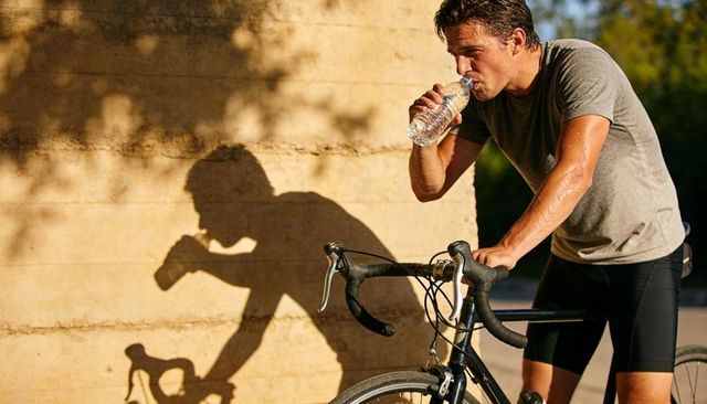 Fit cyclist taking hydration break by sunlit concrete wall with bike shadow at golden hour