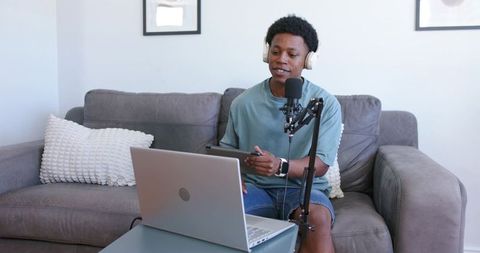 Young black creator recording podcast on sofa with laptop, tablet and mic