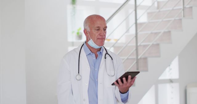 Senior male doctor standing in bright hospital staircase area, using tablet for medical records or research. Stethoscope around neck, wearing a white coat. Suitable for healthcare, medical technology, electronic health records, and modern medical practice themes.