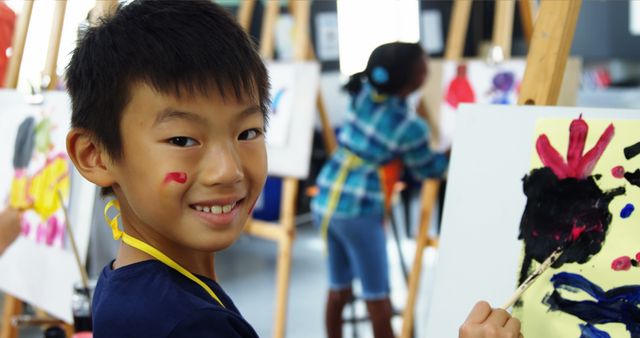 Young boy smiling while painting on canvas in a lively art class. Colorful paints and creativity visible in the background. Ideal for educational materials, children's activities promotions, and art education blogs.