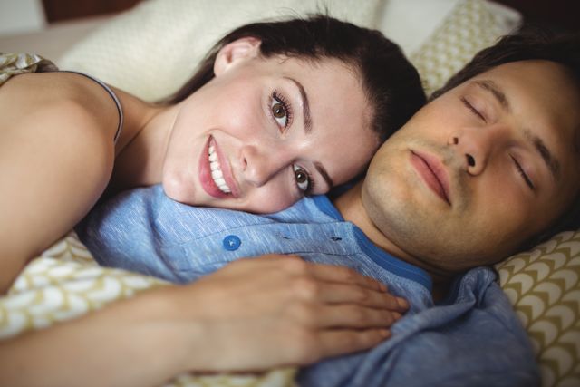 Romantic couple lying in bed, woman smiling while resting her head on man's chest. Ideal for use in relationship blogs, articles about love and intimacy, advertisements for bedding or sleep products, and social media posts celebrating togetherness and romance.