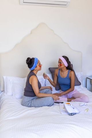 Diverse female friends enjoying at-home spa night applying under-eye gel patches on bed