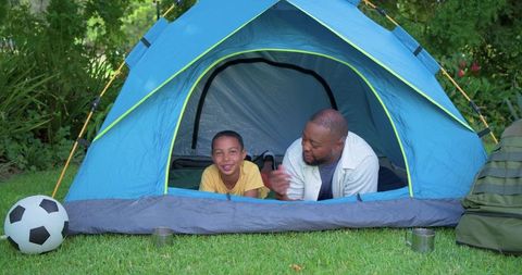African american father and son camping in backyard tent, laughing and talking, family bonding outdo
