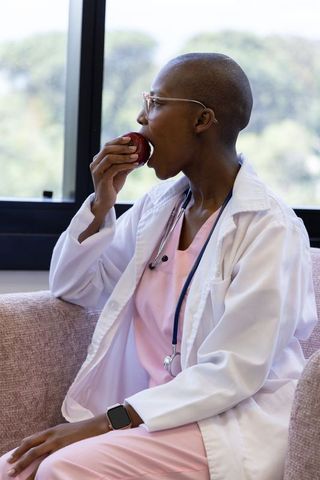 Female clinician eating apple on break in clinic lounge wearing lab coat and stethoscope