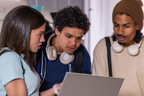 Diverse teens collaborating over laptop in school hallway with headphones and backpacks