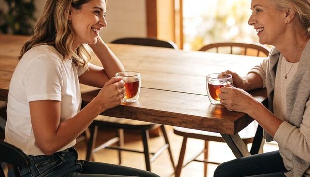 Older woman and younger woman sharing tea and laughing at sunlit wooden table