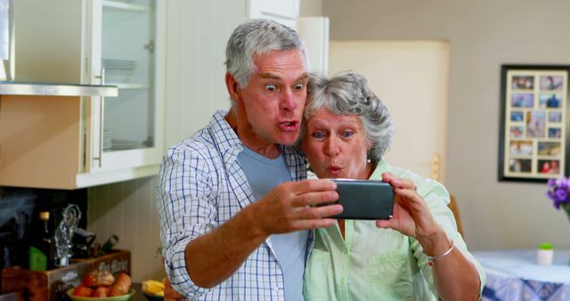 Elderly couple stands in kitchen, taking selfie together, exhibiting excitement and joy. They appear playful and full of love, capturing a cherished moment with a smartphone. This image is ideal for themes related to senior life, family bonds, modern technology usage by older generations, and creating joyful memories.