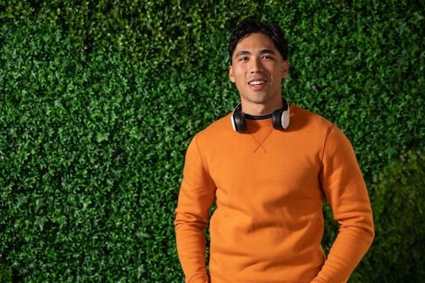 Smiling asian man wearing orange sweater and headphones by green hedge