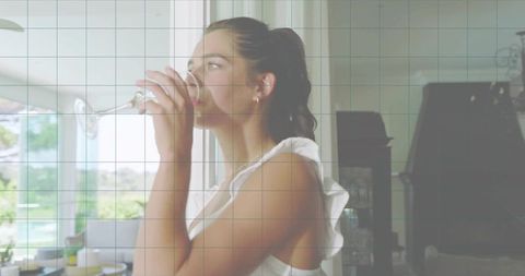 Young woman sipping wine by sunlit window in ruffled white top enjoying elegant morning
