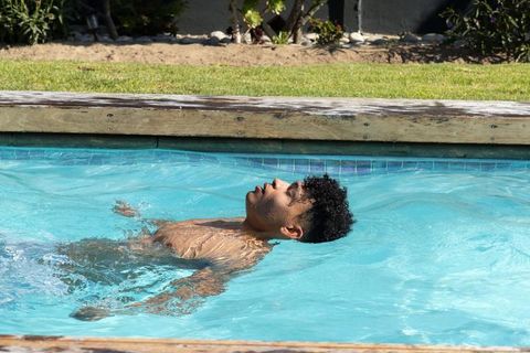 African american man floating and relaxing on back in backyard turquoise pool