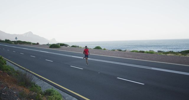 Man jogging on coastal road near ocean during early morning. Ideal for promoting fitness, healthy lifestyle, running gear, outdoor exercise, and motivation for athletes. Suitable for blogs, advertisements, and wellness programs highlighting endurance training and scenic running routes.