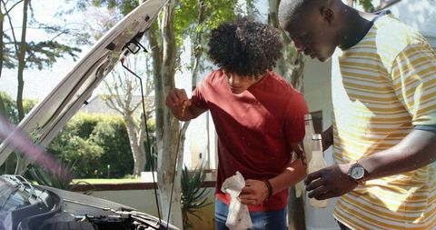 Friends checking car oil dipstick in sunlit suburban driveway
